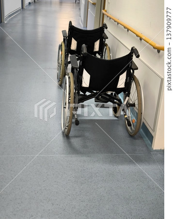 Two empty dark-colored wheelchairs in a bright hospital corridor Two empty dark-colored wheelchairs in a bright hospital corridor 137909777