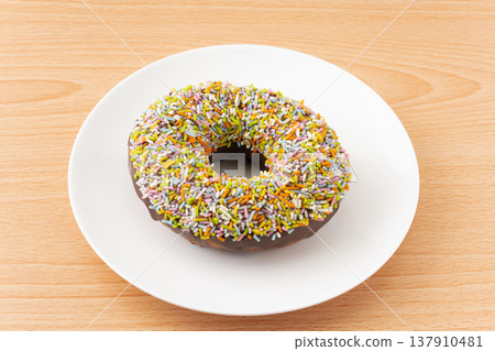 A single chocolate donut sprinkled with colorful sprinkles, placed on a plate. 137910481