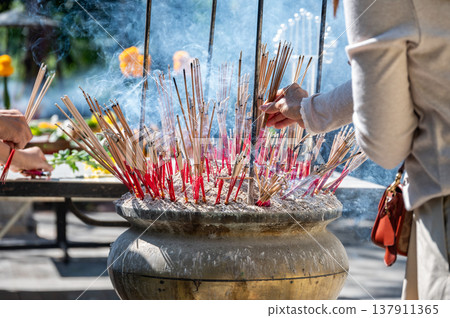 Cropped shot view of woman while place the incense in a pot at Buddhist temple in Thailand. The burned incense may be intended as a symbolic or sacrificial offering to various deities or spirits. Cropped shot view of woman while place the incense in a pot at Buddhist temple in Thailand. The burned incense may be intended as a symbolic or sacrificial offering to various deities or spirits. 137911365
