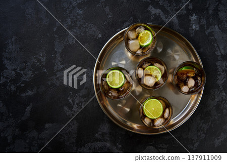 metal tray with cocktail Cuba Libre tumbler over ice and garnished with a lime.Top view. metal tray with cocktail Cuba Libre tumbler over ice and garnished with a lime.Top view. 137911909