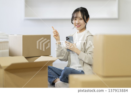 一位微笑的年輕女子坐在被紙箱包圍的座位上(她豎起一根手指),直視鏡頭。畫面是動態的。 一位微笑的年輕女子坐在被紙箱包圍的座位上(她豎起一根手指),直視鏡頭。畫面是動態的。 137913884