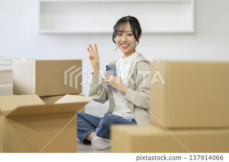 A smiling young woman sits surrounded by cardboard boxes (making a three-finger gesture), looking directly at the camera. The image is of moving. A smiling young woman sits surrounded by cardboard boxes (making a three-finger gesture), looking directly at the camera. The image is of moving. 137914066