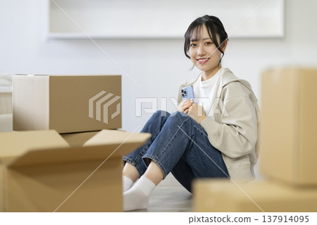 A young woman sits surrounded by cardboard boxes, holding a smartphone, looking directly at the camera. The image evokes the feeling of moving. A young woman sits surrounded by cardboard boxes, holding a smartphone, looking directly at the camera. The image evokes the feeling of moving. 137914095
