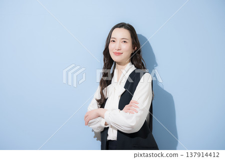 一位留著黑色長髮的時尚女性，身穿商務休閒裝，包括一件長版背心，雙臂交叉抱在胸前。 137914412
