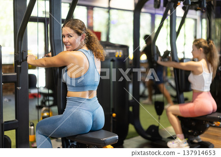 Positive young girl training on chest fly machine in gym Positive young girl training on chest fly machine in gym 137914653