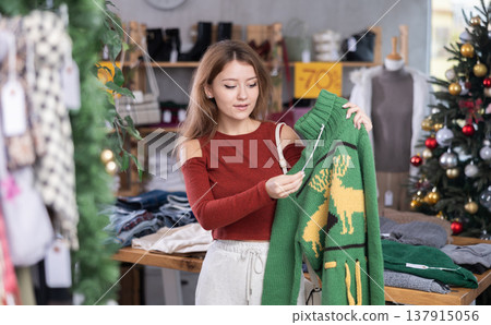 Woman stands in front of a Christmas tree and chooses a green reindeer sweater 137915056