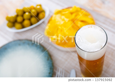 Glass of chilled frothy beer with snacks - potato chips and pickled olives. 137915092