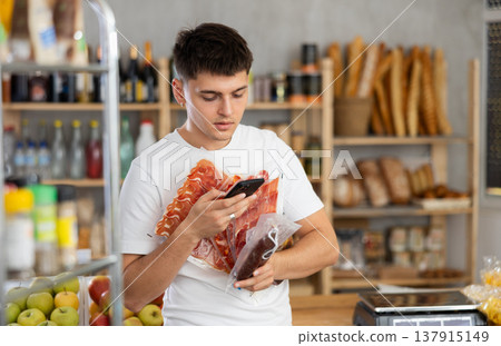 Positive guy purchaser scanning with smartphone QR code of dried sausage or meat in supermarket Positive guy purchaser scanning with smartphone QR code of dried sausage or meat in supermarket 137915149