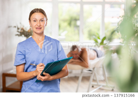 Positive middle-aged masseuse posing with folder in hand in the therapy room Positive middle-aged masseuse posing with folder in hand in the therapy room 137915166