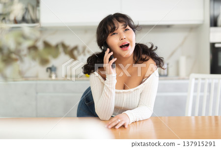 Cheerful girl chatting with friends on phone in home kitchen Cheerful girl chatting with friends on phone in home kitchen 137915291