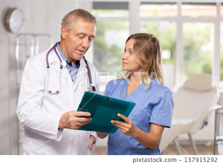 Middle-aged nurse and mature doctor discussing diagnosis standing in cabinet Middle-aged nurse and mature doctor discussing diagnosis standing in cabinet 137915292