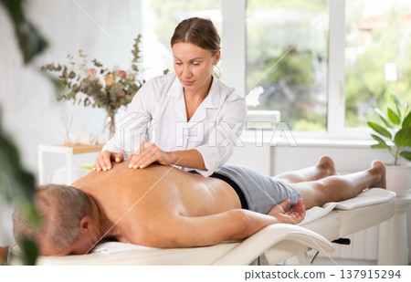 Skilled middle-aged masseuse conducting back massage for man client Skilled middle-aged masseuse conducting back massage for man client 137915294