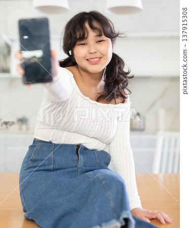 Smiling young girl showing modern smart phone with blank screen, in the living room or in kitchen Smiling young girl showing modern smart phone with blank screen, in the living room or in kitchen 137915308