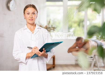 Positive middle-aged masseuse posing with folder in hand in the therapy room Positive middle-aged masseuse posing with folder in hand in the therapy room 137915358