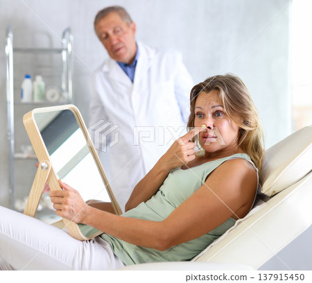 Unsatisfied middle-aged woman holding mirror lying on clinical chair Unsatisfied middle-aged woman holding mirror lying on clinical chair 137915450