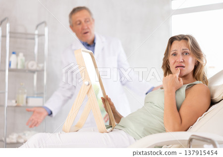 In cosmetology clinic, displeasure client frowns and touches lips, confused doctor in background. 137915454