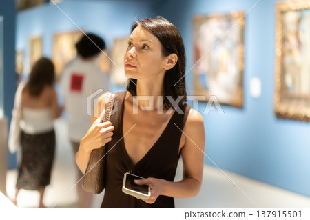 Woman with phone in hands in museum hall, view painting exhibition Woman with phone in hands in museum hall, view painting exhibition 137915501