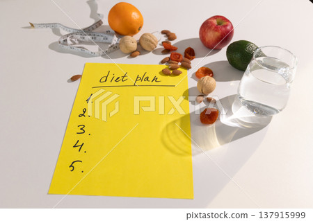 Yellow notepad with empty diet plan outline surrounded by fresh orange, apple, lime, walnuts, almonds, measuring tape, and glass of water. Healthy eating and weight management concept for nutrition Yellow notepad with empty diet plan outline surrounded by fresh orange, apple, lime, walnuts, almonds, measuring tape, and glass of water. Healthy eating and weight management concept for nutrition 137915999