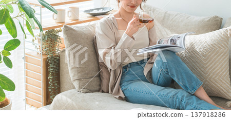 一位穿著時尚的亞洲女性坐在客廳的沙發上，在下午茶時間閱讀雜誌。 137918286