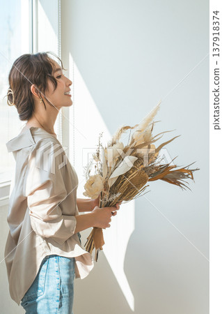 一位衣著時尚的女士手捧乾花,站在客廳窗邊。 一位衣著時尚的女士手捧乾花,站在客廳窗邊。 137918374