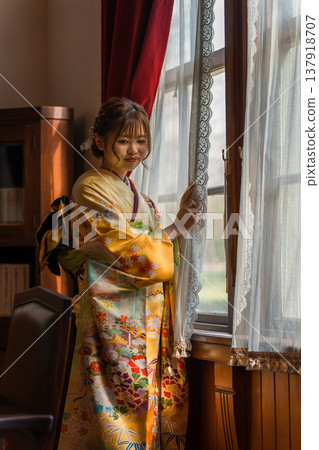 A 20-year-old woman in a furisode kimono gazes out the window of a retro, antique Western-style mansion/studio. 137918707