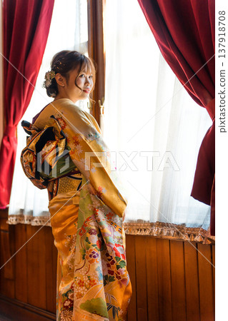 A 20-year-old woman in a furisode kimono gazes out the window of a retro, antique Western-style mansion/studio. 137918708
