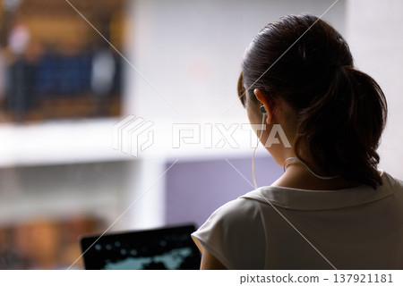 A woman's back as she looks at a PC screen with earphones on. 137921181