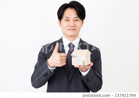 A middle-aged businessman holds up a model of a detached house and gives a thumbs-up. A middle-aged businessman holds up a model of a detached house and gives a thumbs-up. 137921799