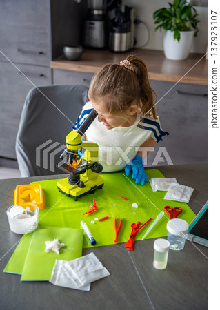 Young girl studying crystal structure through microscope during home science experiment with growing crystals kit. Early STEM education and curiosity driven scientific exploration at home. Young girl studying crystal structure through microscope during home science experiment with growing crystals kit. Early STEM education and curiosity driven scientific exploration at home. 137923107