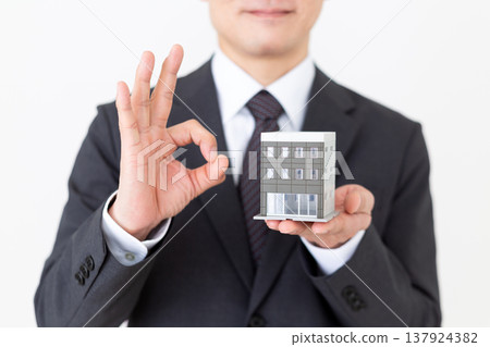 A middle-aged businessman makes a circular sign while holding a model of a residential building in his company building. A middle-aged businessman makes a circular sign while holding a model of a residential building in his company building. 137924382