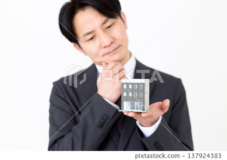 A middle-aged businessman ponders over a residential model of a company building. A middle-aged businessman ponders over a residential model of a company building. 137924383