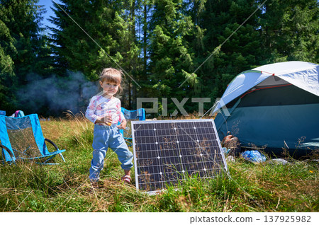Young child playing near tourist tent with photovoltaic solar panel in summer. On background of dense forest. Integration of renewable energy in outdoor camping activities. 137925982