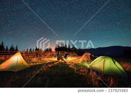 Silhouette of couple hikers resting by campfire under star-studded sky, near illuminated tents at campsite. Night camping in mountains, night sky create magical and peaceful atmosphere. Silhouette of couple hikers resting by campfire under star-studded sky, near illuminated tents at campsite. Night camping in mountains, night sky create magical and peaceful atmosphere. 137925983