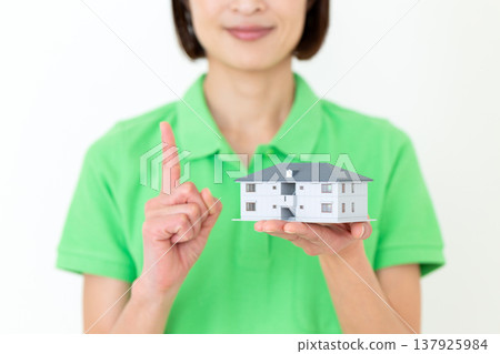A middle-aged female care worker holds up a model house and raises her index finger. A middle-aged female care worker holds up a model house and raises her index finger. 137925984