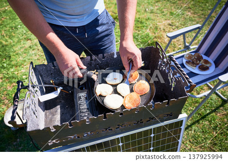 Close up of person cooks golden brown pancakes on griddle over outdoor stove. Solar panel, plate of finished pancakes rests on nearby chair. Scene set on sunny grassy area. Close up of person cooks golden brown pancakes on griddle over outdoor stove. Solar panel, plate of finished pancakes rests on nearby chair. Scene set on sunny grassy area. 137925994