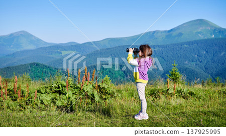 Young girl stands in grassy meadow, looking through binoculars, wearing yellow and purple sweatshirt. Behind her, rolling green hills and mountains stretch into distance under clear blue sky. 137925995