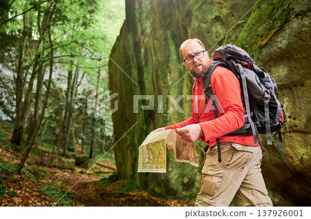 Bearded tourist man studies map while stands in front of large rock in dense forest. Traveler with grey backpack and glasses, planning outdoor adventure, hiking or climbing route. Bearded tourist man studies map while stands in front of large rock in dense forest. Traveler with grey backpack and glasses, planning outdoor adventure, hiking or climbing route. 137926001