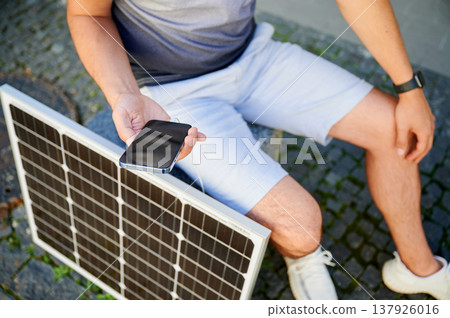 Cropped view of man using smartphone connected to photovoltaic solar panel. Integration of sustainable renewable energy into everyday life, showing practical use of solar power for charging devices. Cropped view of man using smartphone connected to photovoltaic solar panel. Integration of sustainable renewable energy into everyday life, showing practical use of solar power for charging devices. 137926016