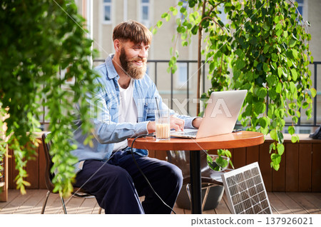 Smiling bearded man works on laptop at outdoor round table, with glass of iced coffee. Solar panel nearby, emphasizing modern eco-friendly workspace that combines technology and sustainability. Smiling bearded man works on laptop at outdoor round table, with glass of iced coffee. Solar panel nearby, emphasizing modern eco-friendly workspace that combines technology and sustainability. 137926021