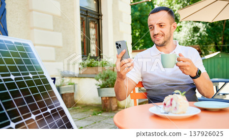 Young man charging smartphone from solar panel while sitting in cafe. Handsome male drinking coffee and checking his phone next to solar battery. Conscious male choosing sustainable lifestyle. Young man charging smartphone from solar panel while sitting in cafe. Handsome male drinking coffee and checking his phone next to solar battery. Conscious male choosing sustainable lifestyle. 137926025