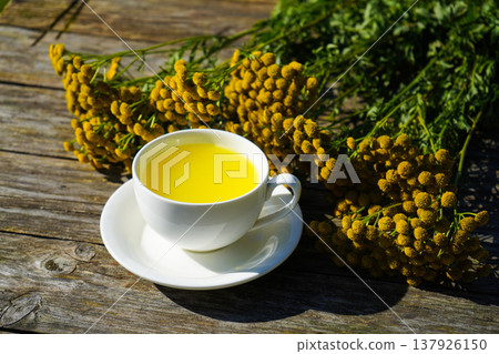 White cup with healthy medicine tansy tea in wooden tray close up. Healthy herbal tansy tea has choleretic and antispasmodic effect. 137926150