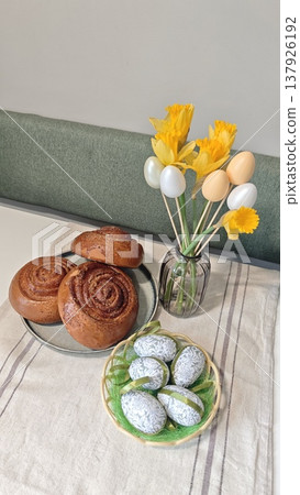 Easter-themed table setting featuring freshly baked cinnamon rolls, decorative eggs, and a bouquet of yellow flowers in a glass vase on a linen tablecloth Easter-themed table setting featuring freshly baked cinnamon rolls, decorative eggs, and a bouquet of yellow flowers in a glass vase on a linen tablecloth 137926192