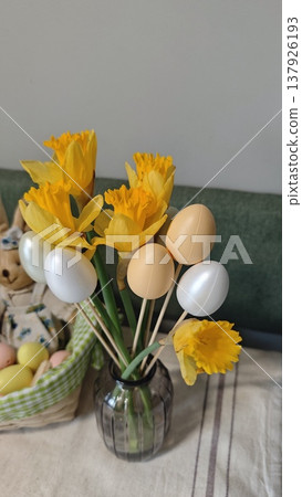 Yellow daffodils and decorative eggs arranged in a glass vase on a table with a basket of pastel-colored eggs in the background for Easter celebration Yellow daffodils and decorative eggs arranged in a glass vase on a table with a basket of pastel-colored eggs in the background for Easter celebration 137926193