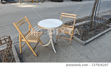 Two wooden chairs and a round table set on a sidewalk near a street, with a decorative metal fence and urban environment visible in the background Two wooden chairs and a round table set on a sidewalk near a street, with a decorative metal fence and urban environment visible in the background 137926207