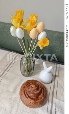 Decorative arrangement featuring yellow daffodils and pastel eggs in a vase alongside a cinnamon roll and a white ceramic bunny on a table with a linen cloth Decorative arrangement featuring yellow daffodils and pastel eggs in a vase alongside a cinnamon roll and a white ceramic bunny on a table with a linen cloth 137926371