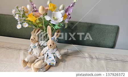Decorative Easter bunnies in floral attire positioned beside a vibrant bouquet of artificial flowers on a neutral tablecloth in a bright indoor setting Decorative Easter bunnies in floral attire positioned beside a vibrant bouquet of artificial flowers on a neutral tablecloth in a bright indoor setting 137926372