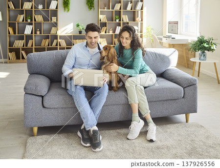 Happy couple with dog using laptop at home on sofa, remote work and leisure Happy couple with dog using laptop at home on sofa, remote work and leisure 137926556