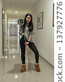 Brunette woman in casual clothes holds studio lighting equipment in hallway with shiny tile floor and neutral walls. 137927776