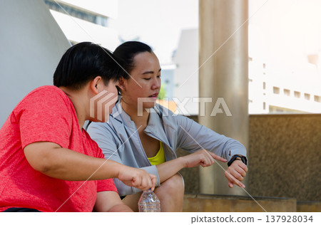 Two Asian women taking a break after outdoor exercise sitting on concrete steps in modern urban city.One holding water bottle and other checking fitness data on smartwatch. Workout and healthy concept Two Asian women taking a break after outdoor exercise sitting on concrete steps in modern urban city.One holding water bottle and other checking fitness data on smartwatch. Workout and healthy concept 137928034