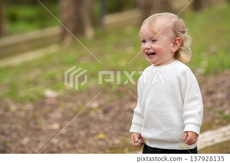 Cute blonde kid playing and smiling outside in a park. Happy kid playing outside on a spring day. Adorable caucasian child walking in a garden and laughing. 137928135